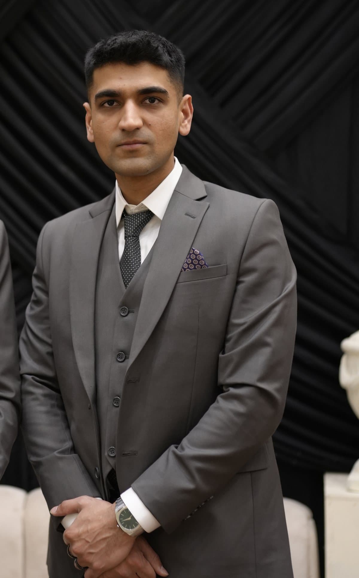 Ali Asif standing in a grey suit and tie against dark drapery